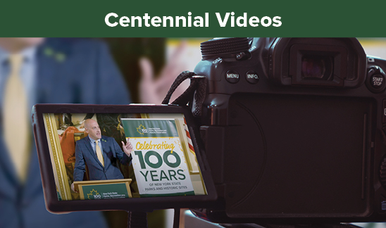 Centennial Videos. A DSLR camera with the live digital viewfinder open. In the viewfinder is Commissioner Pro Tempore Randy Simons speaking at a podium. Over his should is a banner with the text, "Celebrating 100 Years."