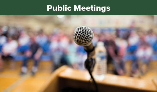 Public Meetings. A microphone at a wooden podium. The perspective of the photograph is from the person speaking, looking out into a crowd of people. There is clearly a large crowd visible in a public meeting space, however the image is blurred with the camera focus being set so only the microphone is clear.